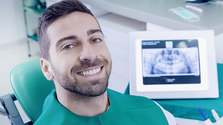 Man in dental chair smiles with dental X-ray screen behind him